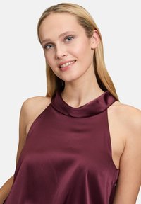 Burgundy sleeveless top in a smooth, textured satin fabric. Features a high, draped neckline and an open shoulder design.
