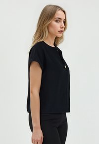 Top nero a maniche corte con scollatura rotonda, caratterizzato da un piccolo dettaglio dorato. Il tessuto sembra liscio e leggermente elasticizzato.