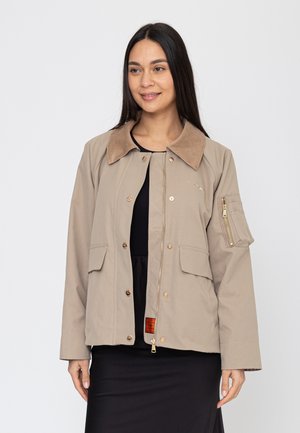 Frau trägt eine beige Jacke mit Cordkragen, vorderen Knöpfen, Reißverschluss und Ärmeltasche über einem schwarzen Kleid vor einem schlichten weißen Hintergrund.