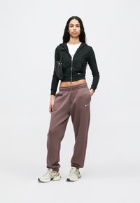 Juodas trumpas zip-up džemperis su kišenėmis, derinamas su laisvomis rožinėmis sportinėmis kelnėmis, akcentuotas mažai juoda tašė ir smėlio spalvos sportbačiais.