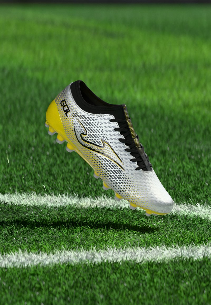 Botín de fútbol blanco y amarillo con cordones y logo negros, reposando sobre un campo de césped verde con líneas de delimitación blancas.