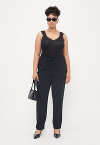 Schwarzer Bodysuit mit Spitzenbesatz, hochtaillierte schwarze Hose mit Bindegürtel, schwarze Handtasche und spitze Absatzschuhe mit Nieten.