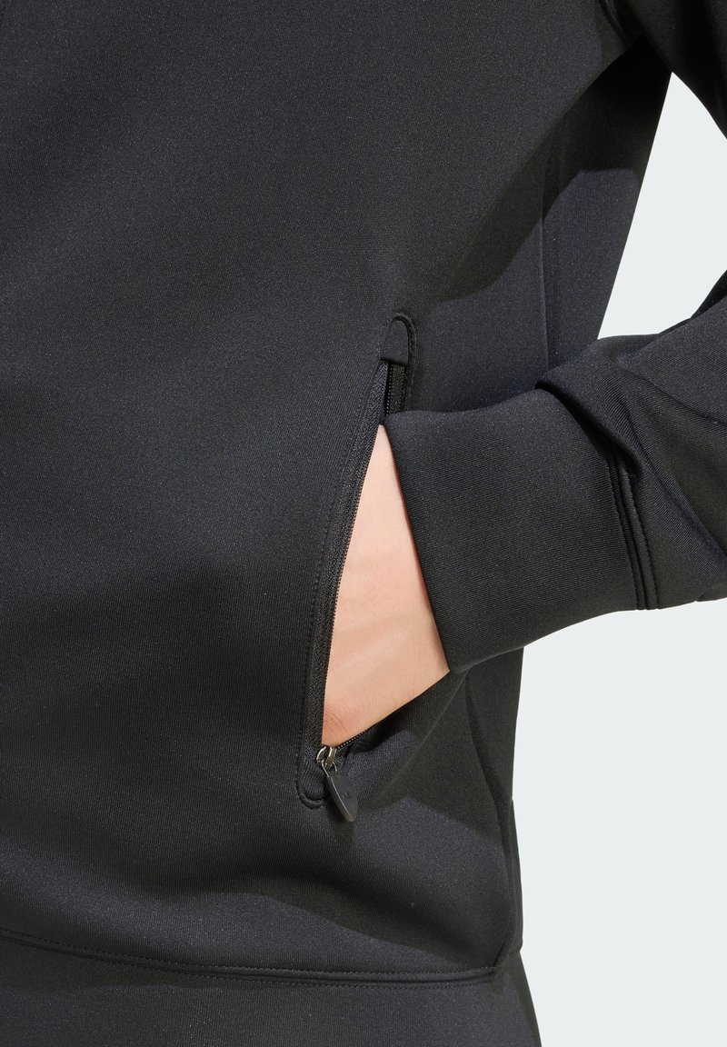 Hand teilweise in einer Reißverschlusstasche eines schwarzen langärmligen Kleidungsstücks mit Bündchen am Handgelenk vor hellem Hintergrund.