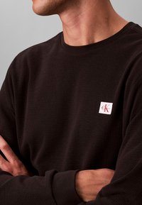 Czarna koszula z długim rękawem wykonana z teksturowanej tkaniny, z małą białą naszywką z logo Calvin Klein na piersi i ściągaczami przy rękawach.