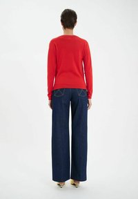 Un pull tricoté rouge à col rond associé à un jean en denim foncé à jambes larges avec des détails de couture sur les poches arrière. Des talons jaunes viennent compléter le look.