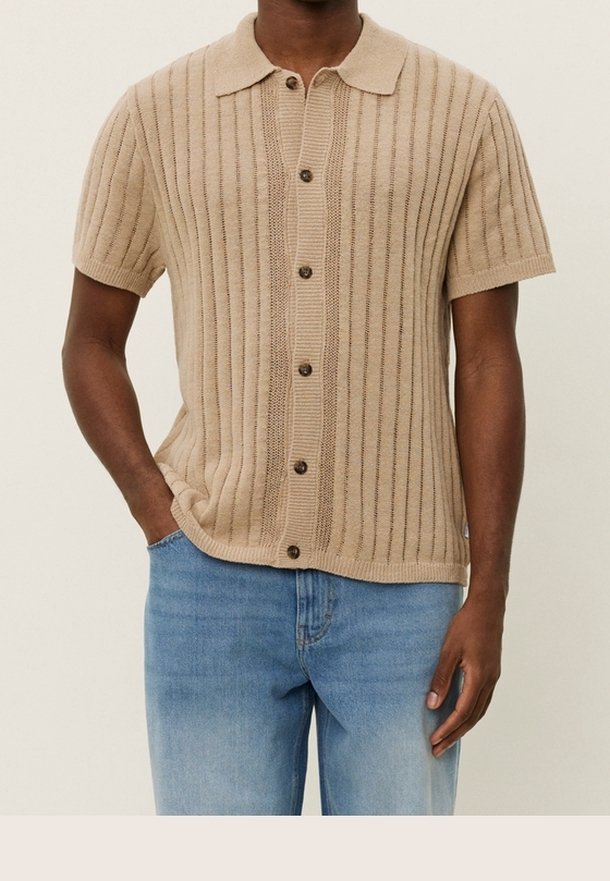 Beżowa, dziana koszulka polo z krótkim rękawem o pionowej, ryżowej fakturze, z zapięciem na guziki i miękkim kołnierzykiem. Nosi ją z jasnoniebieskimi dżinsami.