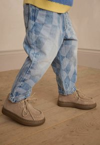 Beiga mockaskor med snörning, ljusbruna gummisålar. Blå mönstrade denimjeans med avslappnad passform. Bakgrund av trägolv.