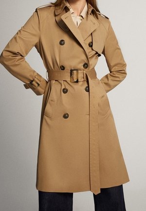 Woman wearing a knee-length beige trench coat with double-breasted buttons and a belted waist, standing with hands on hips.
