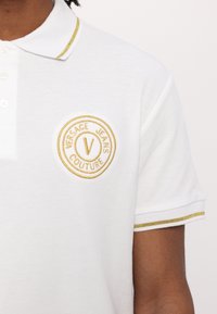 Biała koszulka polo z teksturowanej tkaniny, z okrągłym złotym haftowanym logo na piersi, które brzmi "VERSACE JEANS COUTURE". Złote akcenty wykańczają kołnierz.