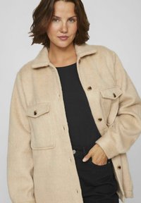 Beige villasekoitteinen takki, jossa on napillinen etuosa, kaulus ja kaksi rintataskua. Pehmeä tekstuuri, rento istuvuus ja ruskeat napit korostavat muotoilua.