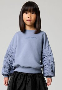 Sudadera azul claro con mangas fruncidas y puños acanalados, con "ALLSAINTS" bordado al frente, combinada con una falda negra de tul.