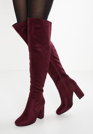 Bourgogne ruskindsstøvler til knæet med blokhæl, båret med sorte strømpebukser på krydsede ben mod en hvid baggrund.