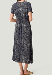 Femme portant une robe midi à motif bleu marine avec des manches courtes et un détail de dos en forme de clé, associée à des sandales slip-on beiges.