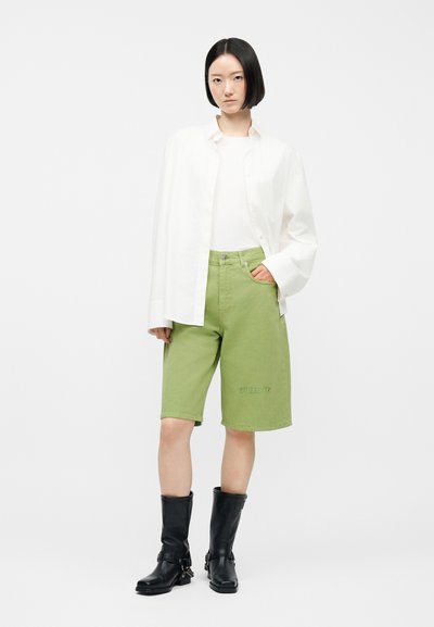 Noor naine seisab käega taskus, seljas ülikasukas valge särk, valge top, rohelised põlveni püksid ja mustad keskmise säärega saapad.