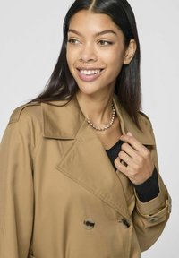 Brun dubbelknäppt trenchcoat med stora slag, knappdetaljer och en mjuk textur, i kombination med en svart topp och pärlhalsband.