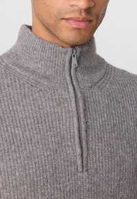Grå zip-up genser med høy krage, med en teksturert ribbestrikket mønster og en sølvglinsende glidelås. Myk stoff og tettsittende design.
