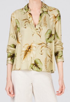 Beige blouse met lange mouwen met een groen-bruine tropische bladprint, gedragen met lichtgekleurde broek.