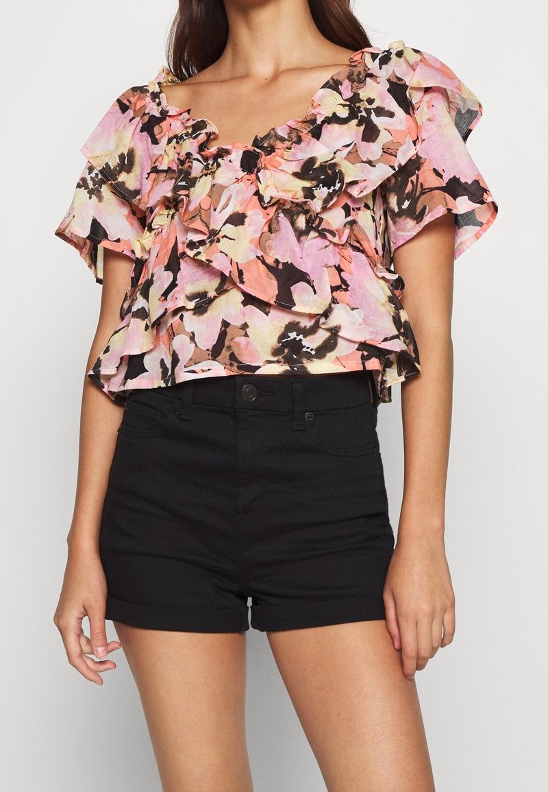 Blomsterblusen har flæser på ærmerne, en V-udskæring og en farveskala i lys pink, sort og beige. Den er parret med sorte højtaljede shorts.