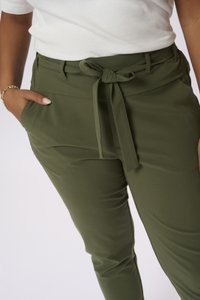 Pantalons extensibles vert olive à taille haute, avec un nœud noué à l'avant et des poches latérales. Texture lisse et coupe ajustée.