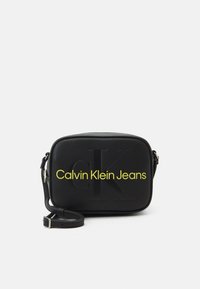 Borsa a tracolla nera realizzata in morbida ecopelle, con un logo impresso e un testo giallo brillante "Calvin Klein Jeans". Cinghia regolabile.