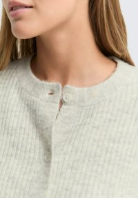 Prim-plan al unei femei îmbrăcate cu un cardigan cu dungi gri deschis, cu guler rotund, fixat cu două butoane mici albe.