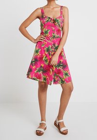 Robe d'été florale avec une base rose, comportant des feuilles vertes et des fleurs jaunes, décolleté carré et bretelles ajustables. Associée à des sandales blanches.