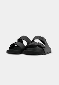 Une paire de sandales noires texturées avec deux sangles à boucle réglables et des semelles nervurées, présentées sur un fond clair et uni.