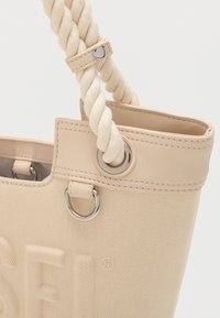 Μπεζ τσάντα ώμου από καμβά με διπλές λαβές από στριμμένο σχοινί που είναι προσαρτημένες μέσω μεταλλικών ματιών και ραφή λεπτομέρειας στην πάνω άκρη.