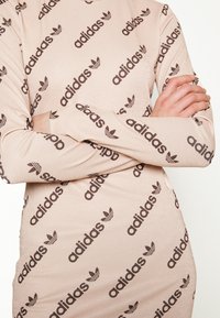 Strak aansluitende jurk met lange mouwen van zacht blush stof met een glanzende afwerking, voorzien van herhalende donkerbruine "adidas"-logo's en trefoil-accenten.
