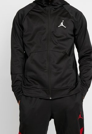 Osoba w czarnej bluzie na zamek z kapturem i czarnych spodniach z białymi i czerwonymi logo Jumpman, ręce w kieszeniach.