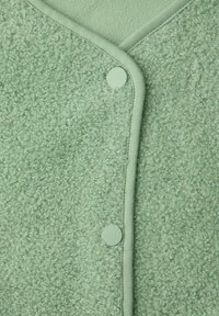 Close-up van lichtgroene, getextureerde stof met twee bijpassende ronde drukknopen en gedetailleerde stoffen afwerking.