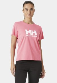 Mujer con el cabello recogido, vestida con una camiseta rosa de Helly Hansen y pantalones negros, de pie contra un fondo blanco liso.
