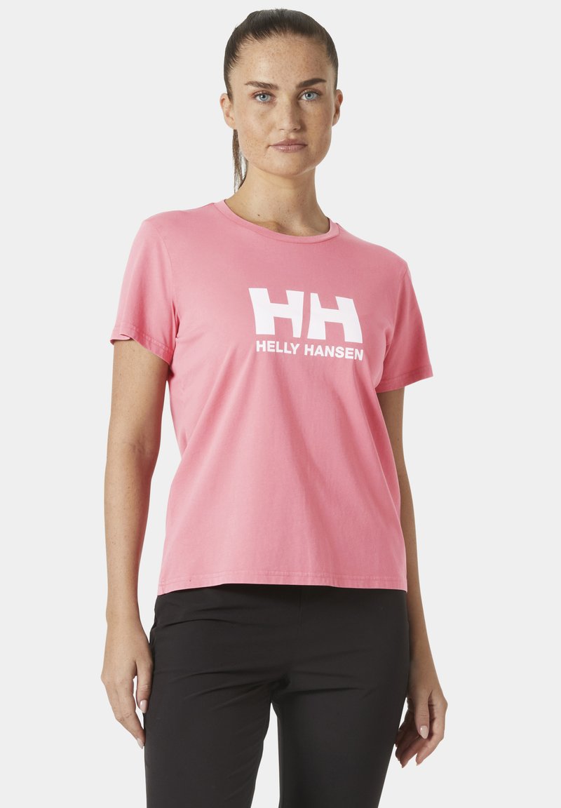 Mujer con el cabello recogido, vestida con una camiseta rosa de Helly Hansen y pantalones negros, de pie contra un fondo blanco liso.