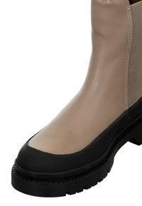 Beige leather ankle boot with black rubber sole, rounded toe, and contrasting stitching detail. Features smooth texture and elastic side panel.