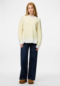 Kreminės ir geltonos spalvos megztinis su pintu raštu, plačiu iškirpimu ir ribbingu apačioje, dera su tamsiai mėlynais, plačios kojų linijos džinsais ir ruda platformų avalyne.