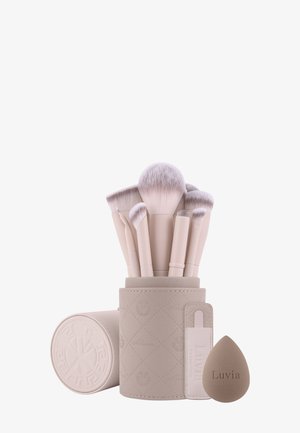 Zestaw siedmiu beżowych pędzli do makijażu w teksturowanym uchwycie, z dopasowaną gąbką do makijażu obok nich.