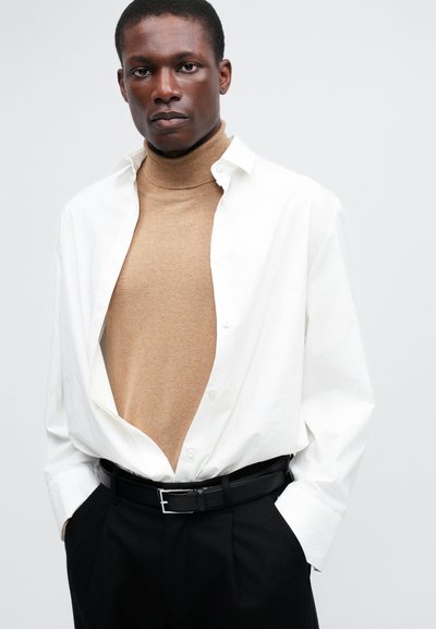 Camisa branca de botão sobre um suéter bege de gola alta, combinada com calças pretas e um cinto preto. Apresenta texturas suaves e um design em camadas.
