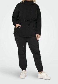 Svart, lätt jacka med dragsko i midjan och fickor, tillsammans med matchande svarta cargobyxor och vita sneakers.