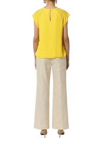 Żółta bluzka z krótkim rękawem i dekoltem w kształcie łezki, zestawiona z jasnymi szerokimi spodniami oraz beżowymi butami na obcasie.