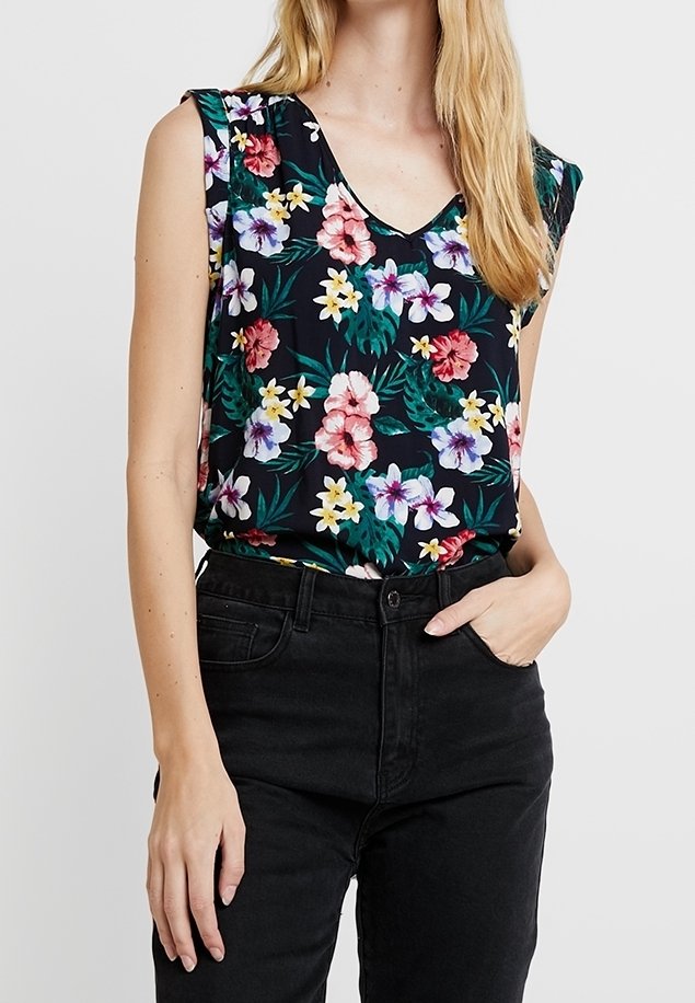 Haut noir sans manches à fleurs roses, violettes et blanches, porté rentré dans un jean noir taille haute, main dans la poche.