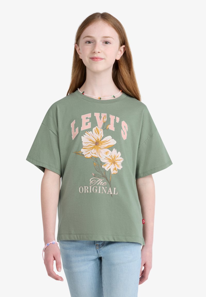 Νέο άτομο με μακριά ανοιχτά καστανά μαλλιά που φοράει πράσινο τ-shirt Levi's με κίτρινα λουλούδια και ανοιχτό μπλε τζιν, στέκεται μπροστά σε λευκό φόντο.
