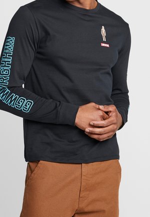 Homme portant une chemise noire à manches longues Levi's avec un texte bleu clair sur la manche gauche et un pantalon marron, les mains croisées à la taille.