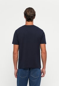 Bărbat cu părul scurt castaniu, îmbrăcat cu un tricou simplu de culoare bleumarin închis și blugi, întors cu spatele pe un fundal alb.