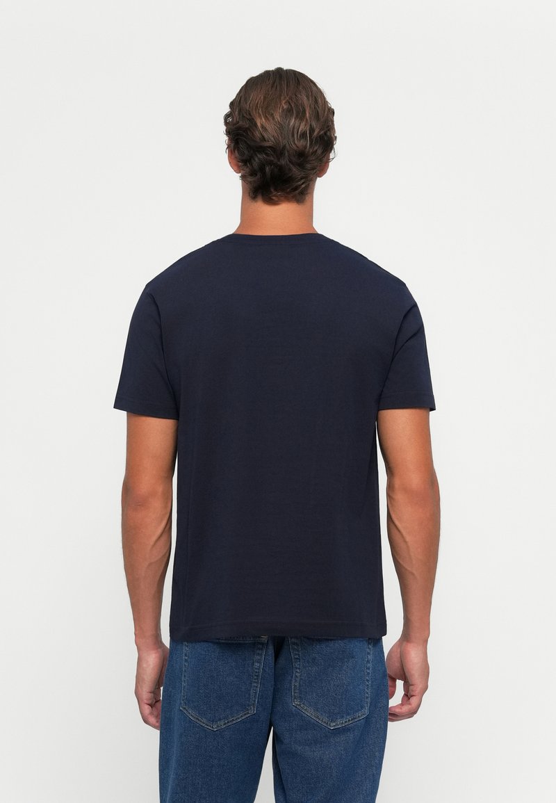 Mężczyzna z krótkimi brązowymi włosami, ubrany w prostą ciemnonawiwową koszulkę i niebieskie dżinsy, zwrócony tyłem na białym tle.