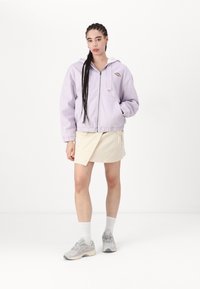 Fialová mikina na zips s bielou fleecovou kapucňou, s malým oranžovým logom. Skombinovaná s béžovou obálkovou sukňou a sivými športovými topánkami.