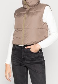 Bezruká péřová vesta světle hnědé barvy s vysokým límcem a plným předním zipem, vyrobená z hladkého lesklého materiálu, kombinovaná s tmavými džíny.