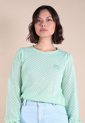 Chemise à manches longues verte menthe avec un motif en diamant, un tissu texturé et un petit patch de logo. Associée à un jean bleu clair.