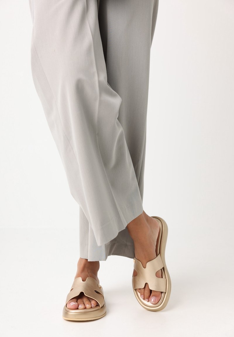 Personne portant des sandales ouvertes beige clair avec un design découpé et un pantalon ample gris clair, debout sur un fond blanc uni.