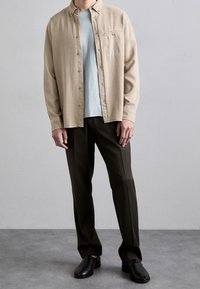 Beige nappipaita vaaleansinisen t-paidan päällä, yhdistetty tummiin housuihin ja mustiin kenkiin. Sileä puuvillatekstuuri, jossa on rintataskun yksityiskohta.