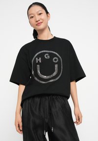 Camiseta de algodón negra con un gran gráfico de purpurina plateada de una carita sonriente con el texto "HGO" dentro, corte relajado y mangas cortas.